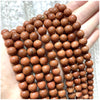 A hand holds a strand of small, round, reddish-brown natural stone beads for jewelry making.