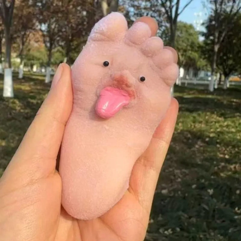 A hand holds a pink silicone fidget toy shaped like a foot with a tongue sticking out in an outdoor setting.