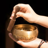 A hand holds a handcrafted brass Tibetan singing bowl and a wooden mallet against a dark background.
