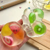 A hand holds a clear squishy stress ball shaped like a fruit, featuring pink and green segments.