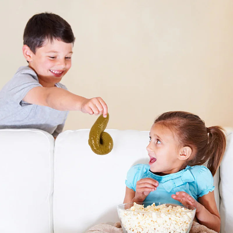 A young boy is playfully putting a soft, green fake poop toy onto a young girl's face while she watches.