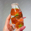 A hand holds a clear glass bottle filled with orange fruit pieces, serving as a drink cup toy.