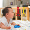Ein Baby spielt mit blauen Entwicklungsringen auf einer weißen Decke in einem hellen Innenraum.
