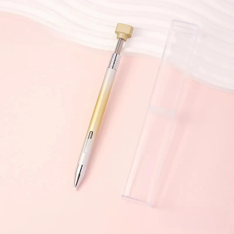 A gold and silver refillable diamond picking pen with a rotating wax tip resting on a pink surface.