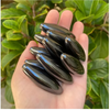 A hand holds several smooth, dark olive green magnetic snake egg fidget toys against a blurred green leaf background.