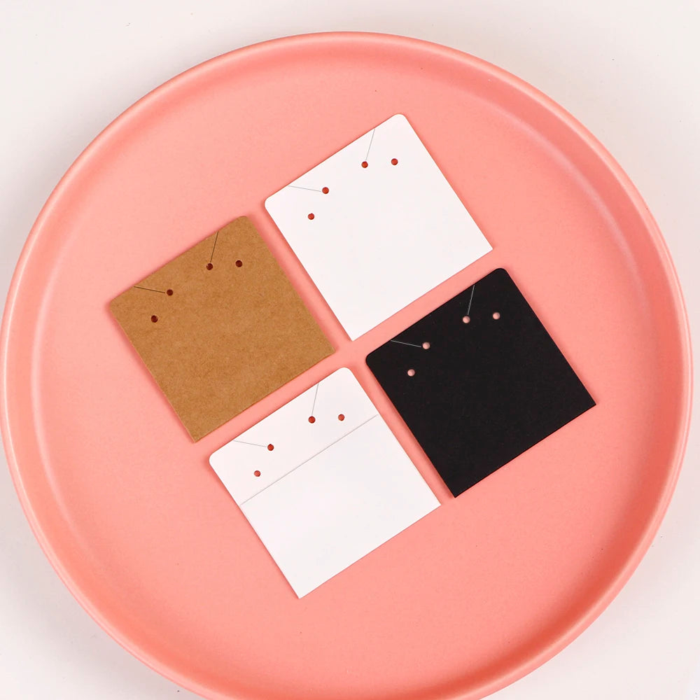 Five square jewelry display cards in kraft brown, white, and black are arranged on a pink plate.
