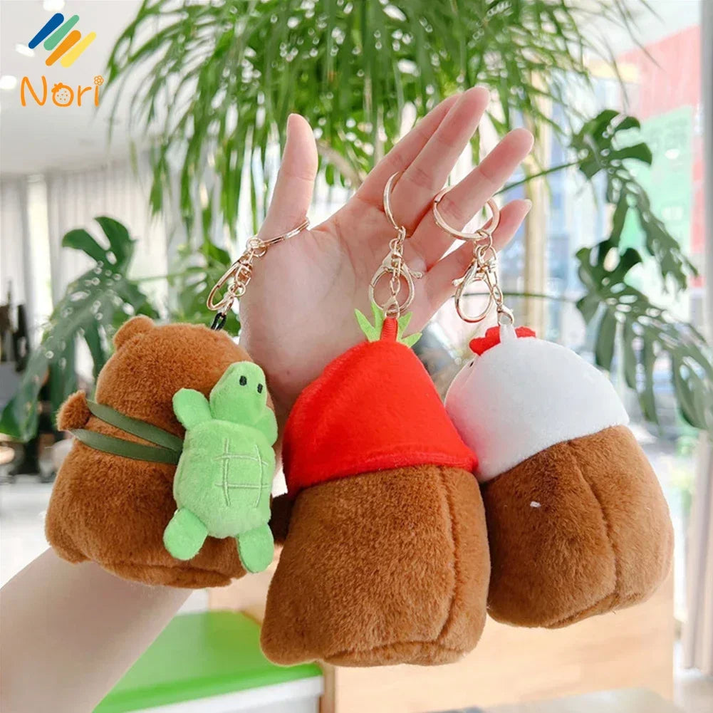 A hand holds three adorable capybara plush keychain charms in shades of brown, green, and white.
