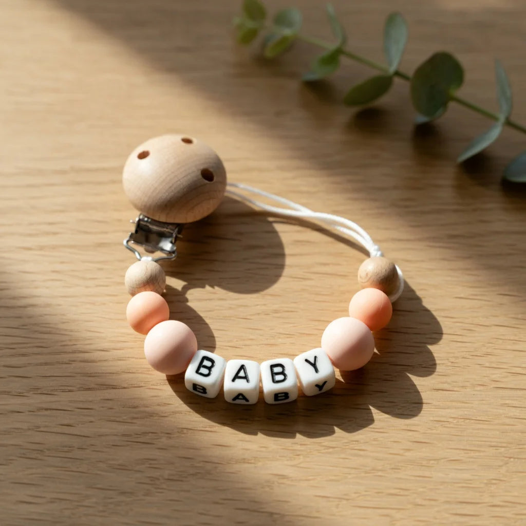 A silicone pacifier clip with a wooden charm shaped like a baby, resting on a light wood surface.