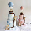 A tiered cake display featuring blue and pink fondant elements, including bear figures and clouds.