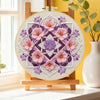 A circular flower embroidery design featuring pink and purple flowers on a beige fabric, displayed on a wooden easel.