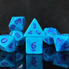 A collection of seven polyhedral dice in shades of blue and purple are displayed on a reflective surface.