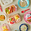 A collection of miniature food items and a pink plastic lunch box with a handle on a light textured surface.
