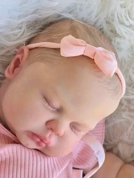 A close-up of a lifelike reborn baby doll with hand-painted skin wearing a pink outfit and headband.