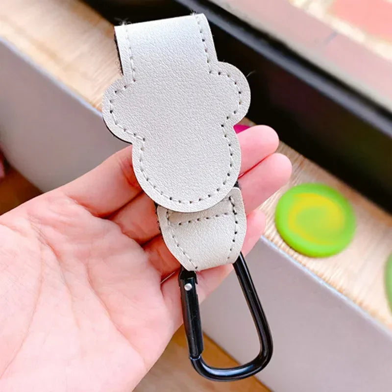 A hand holds a light gray PU leather stroller organizer hook with a black clip.