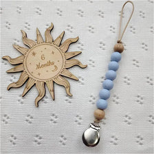 A wooden sun-shaped bead pacifier clip with six blue beads and a silver silicone teether on a light background.