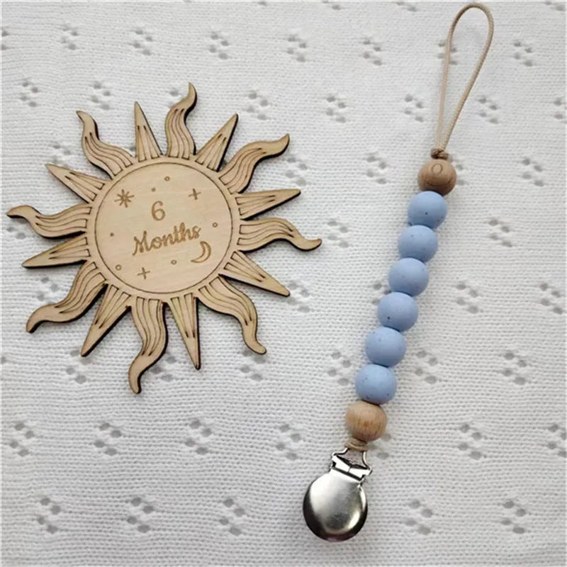 A wooden sun-shaped bead pacifier clip with six blue beads and a silver silicone teether on a light background.