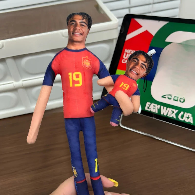 A male football star stuffed doll wearing a red and blue jersey is held by a hand in front of a soccer field background.