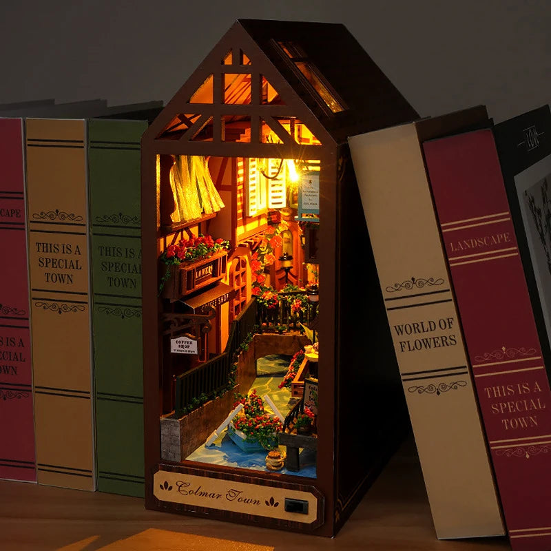 A DIY book nook kit featuring a wooden house structure with warm LED lighting and detailed scenery.