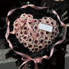 A heart-shaped arrangement of dried pink and beige roses tied with a pink ribbon, presented in black paper.