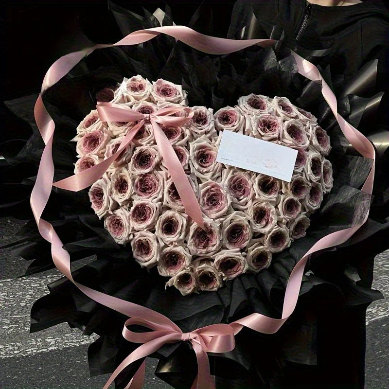 A heart-shaped arrangement of dried pink and beige roses tied with a pink ribbon, presented in black paper.