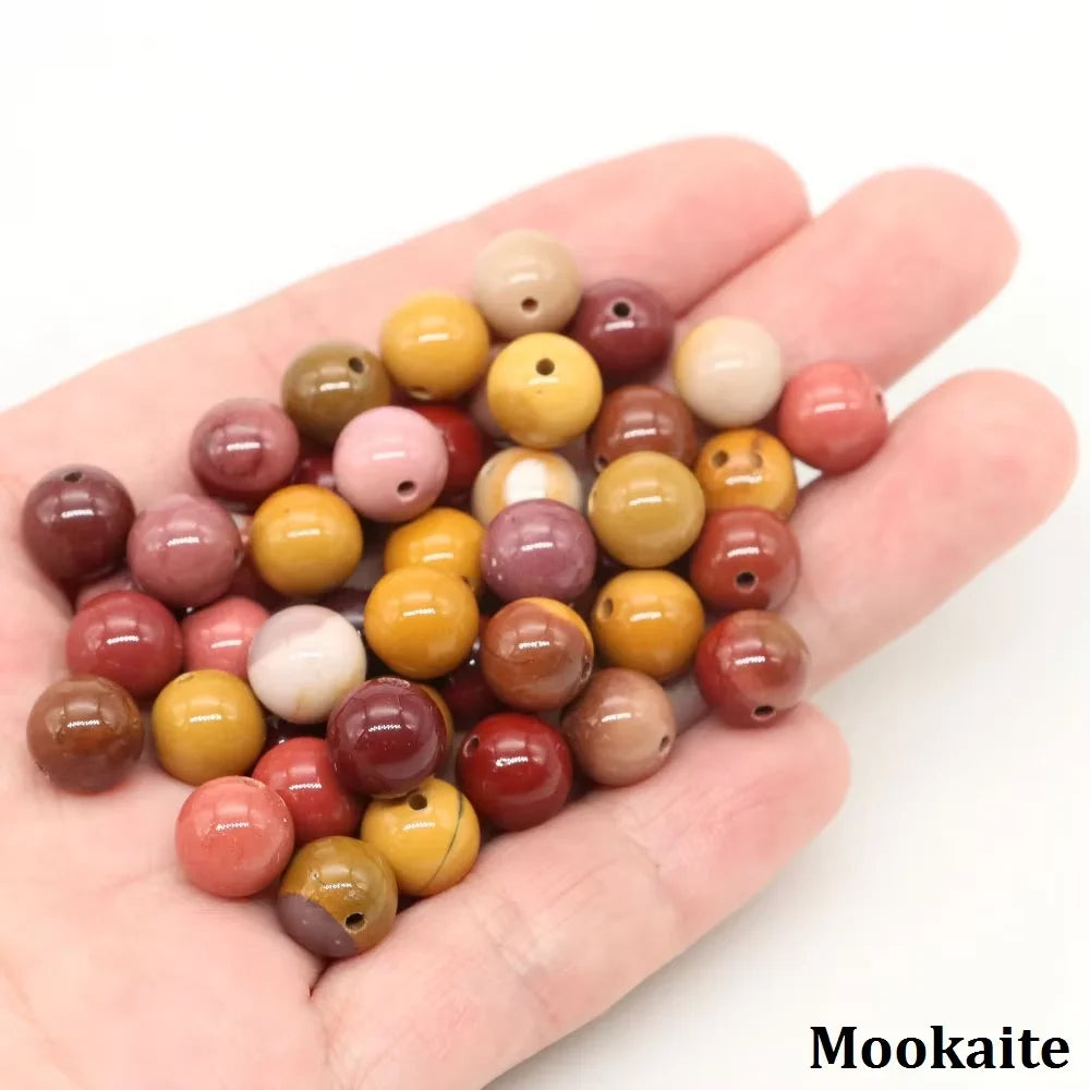A hand holds a collection of natural stone round beads in various shades of pink, red, and yellow for jewelry making.