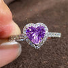 A close-up of a heart-shaped ring featuring a large purple gemstone surrounded by small clear stones.
