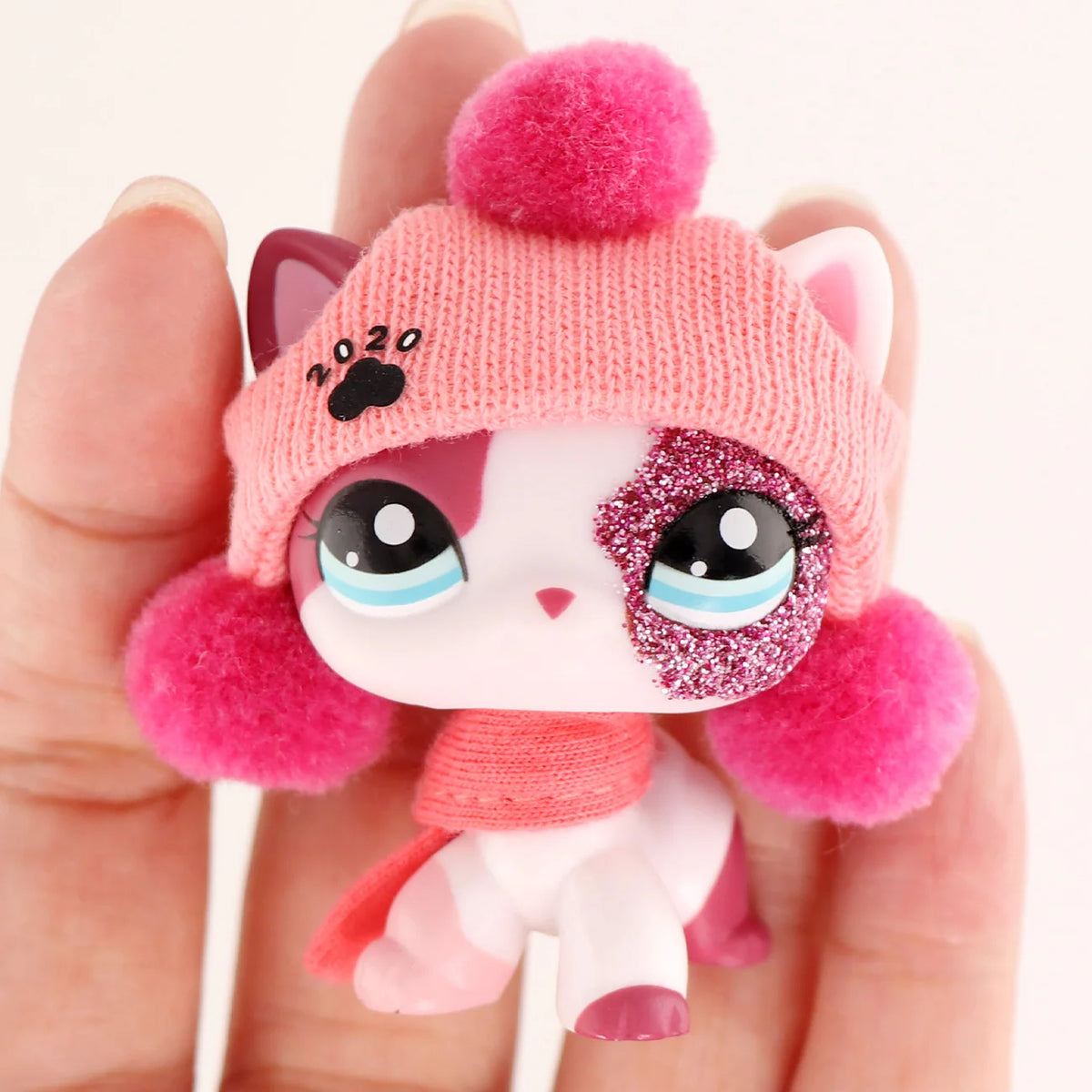A small pink knitted animal figure resembling a cat with pink and white fur and large sparkly eyes is held in a hand.