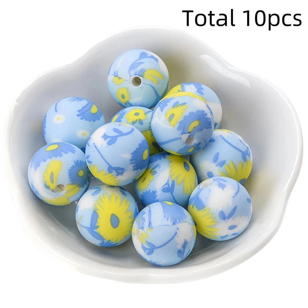 A collection of ten blue silicone beads with yellow floral patterns in a white dish.