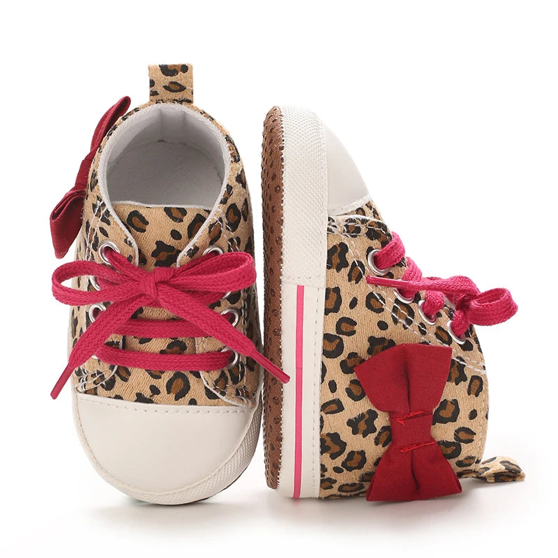 A pair of leopard print soft sole baby sneakers with pink laces and red bows.