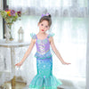 A young girl wearing a turquoise and purple mermaid fantasy dress with a ruffled fishtail skirt standing indoors.