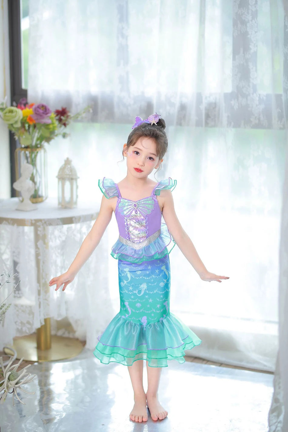 A young girl wearing a turquoise and purple mermaid fantasy dress with a ruffled fishtail skirt standing indoors.