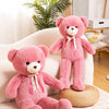 A pink giant plush teddy bear sitting on a woven mat next to a white sofa.