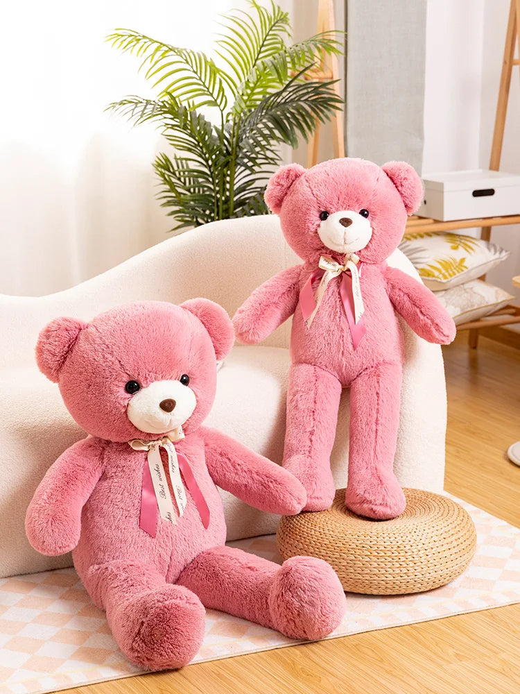 A pink giant plush teddy bear sitting on a woven mat next to a white sofa.