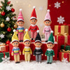 A set of seven elf figurines in various holiday outfits standing on a white surface with Christmas decorations.