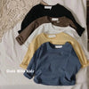 A stack of four kids' solid color long sleeve cotton tees in black, brown, beige, and blue laid on a white surface.