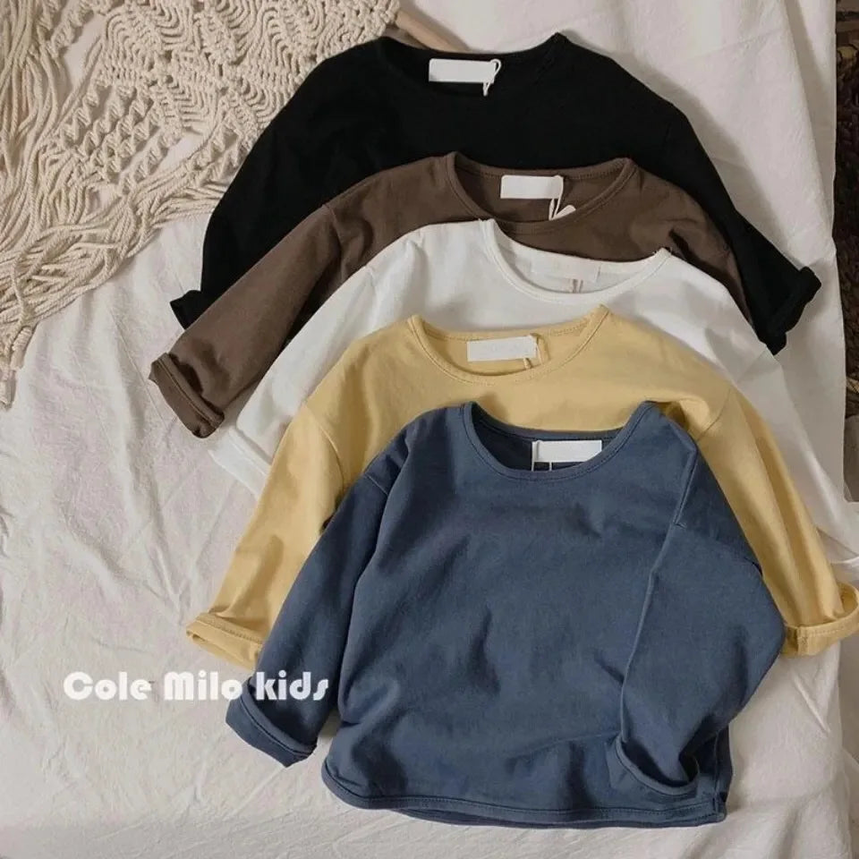 A stack of four kids' solid color long sleeve cotton tees in black, brown, beige, and blue laid on a white surface.