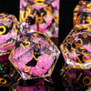 A close-up of a set of pink and gold liquid core dice, shaped like pyramids, reflecting on a dark surface.