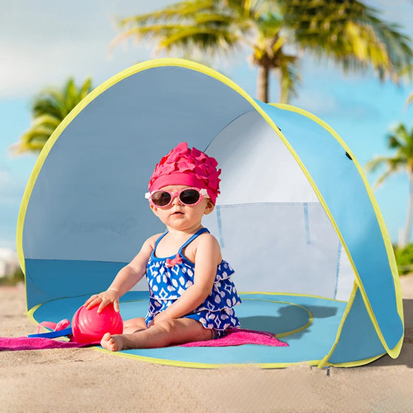 A baby wearing sunglasses and a pink hat sits inside a blue instant pop-up beach tent on the sand.