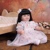 A lifelike reborn baby doll with soft cloth body wearing a white floral dress sitting on a wooden surface.