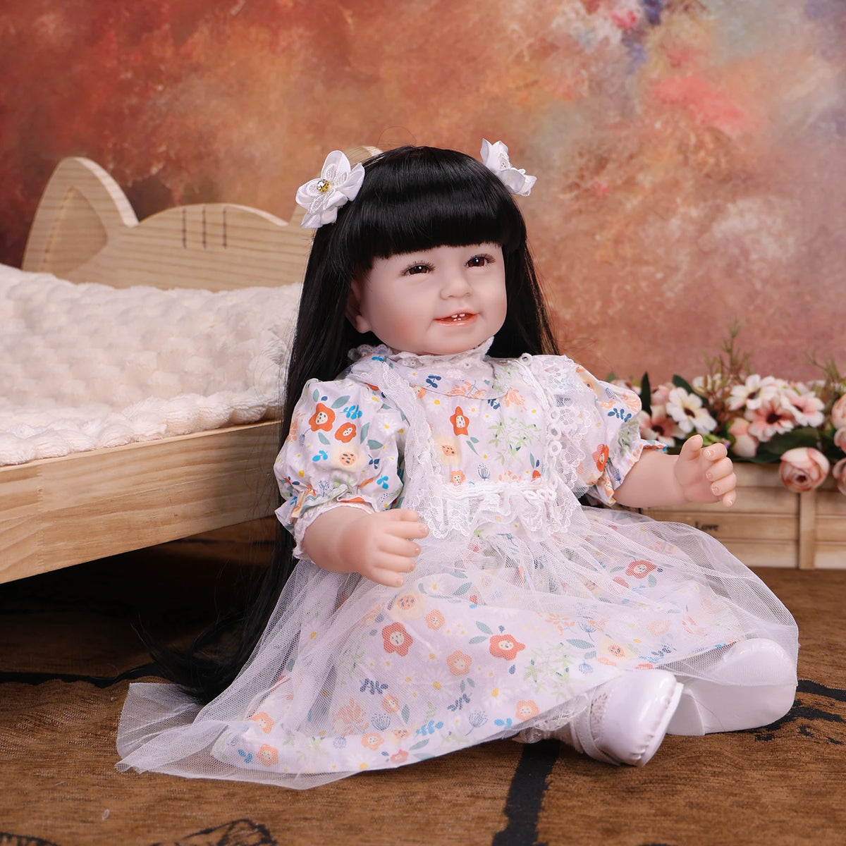 A lifelike reborn baby doll with soft cloth body wearing a white floral dress sitting on a wooden surface.