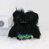 A black, fuzzy monster plush toy with large green eyes and a bright blue patterned collar is shown on a white surface.