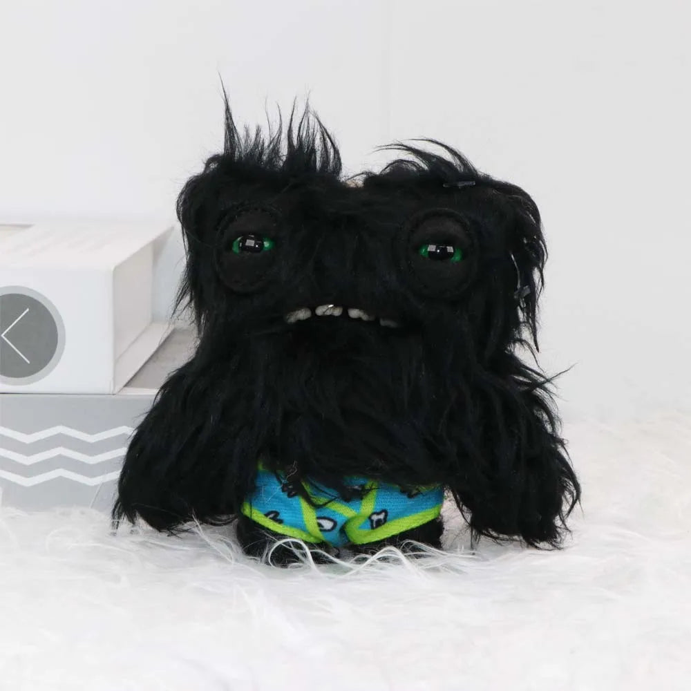 A black, fuzzy monster plush toy with large green eyes and a bright blue patterned collar is shown on a white surface.