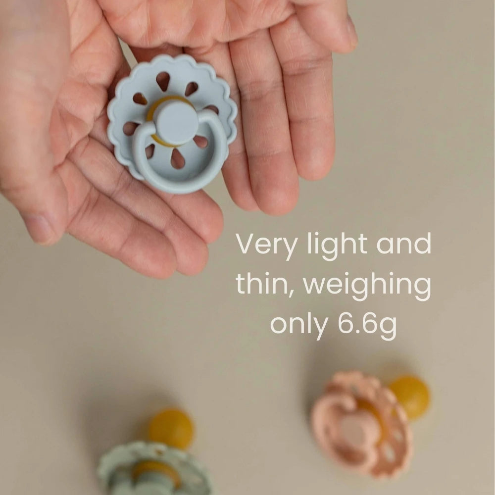 A hand holds a light gray natural latex baby pacifier with a flower-like design.