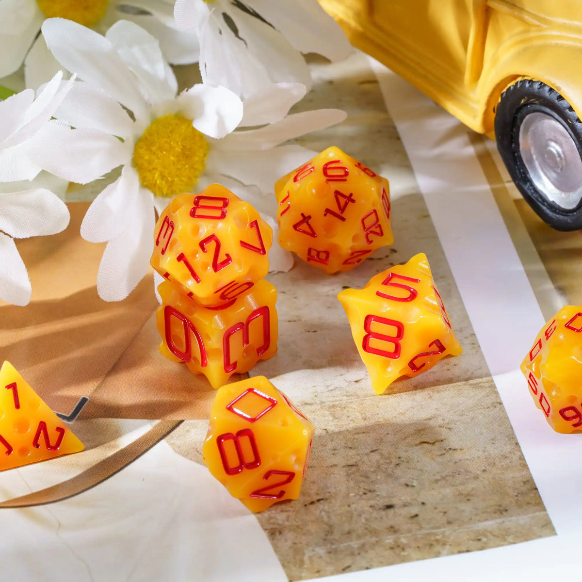 A set of seven yellow and red polyhedral dice resting on a light brown surface next to a white flower.