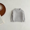 A light gray knit baby pullover sweater lying flat on a white surface next to a wooden plate and a book.