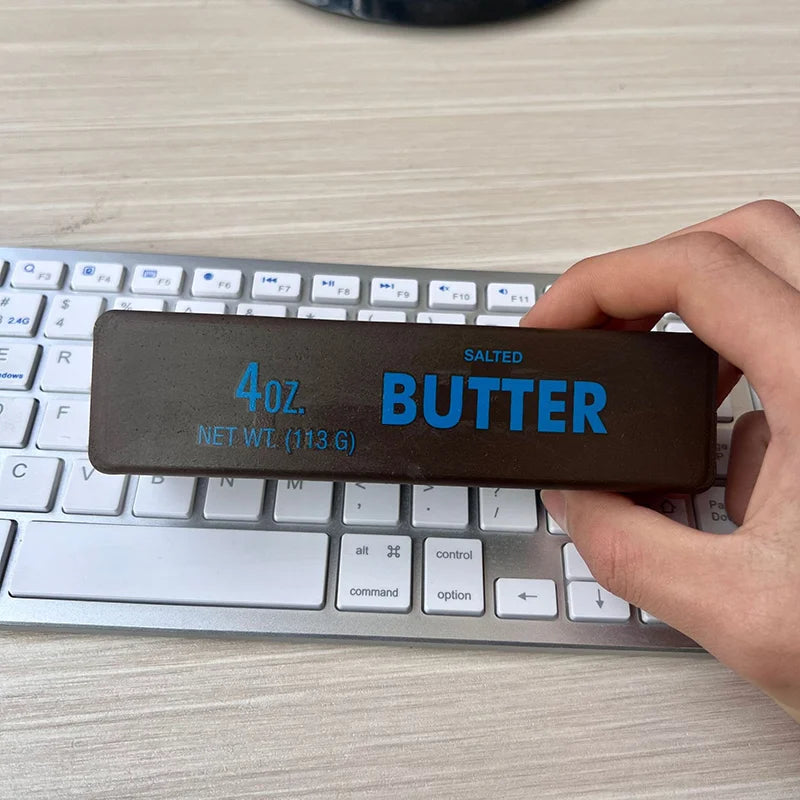 A hand holds a salted butter stick fidget toy with the label 4oz butter on a silver laptop keyboard.