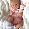 A baby doll wearing a pink outfit and sitting in white fluffy material with a pink rabbit plush toy.
