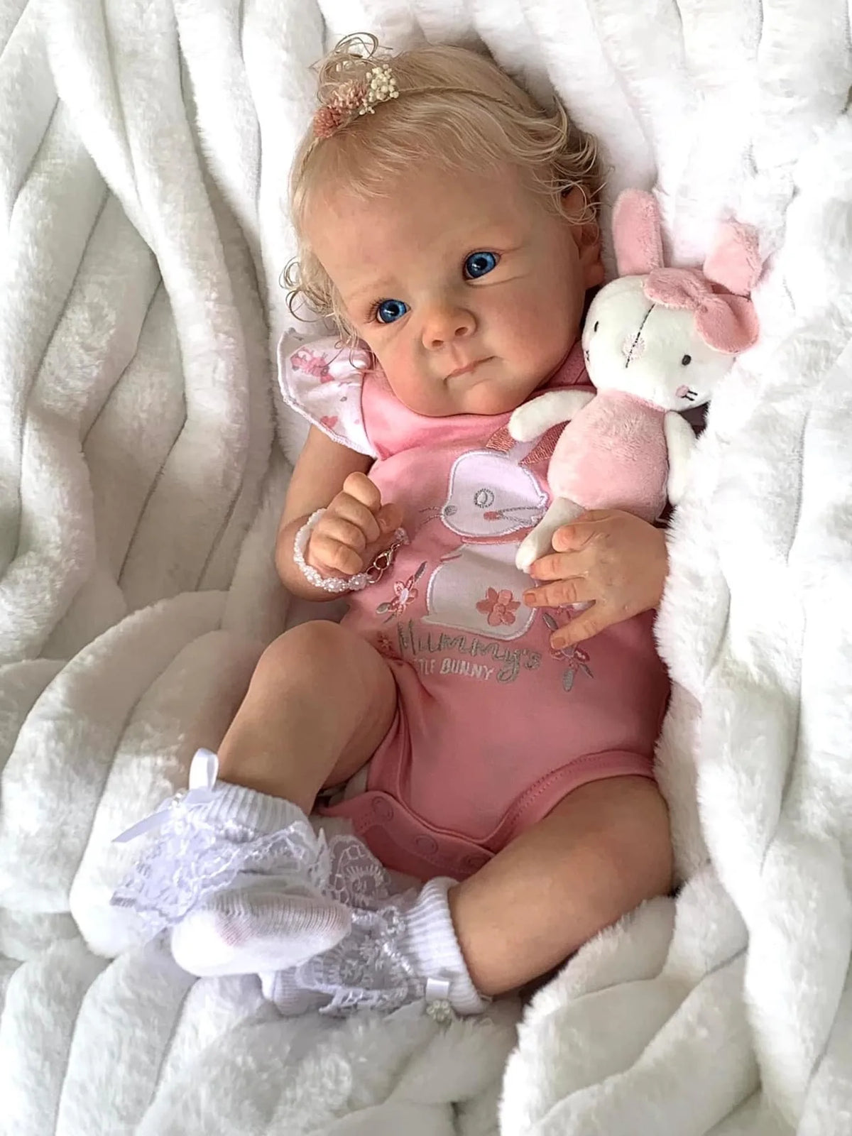 A baby doll wearing a pink outfit and sitting in white fluffy material with a pink rabbit plush toy.