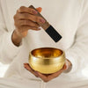 A hand holds a brass singing bowl and a wooden mallet, ready for meditation or yoga.