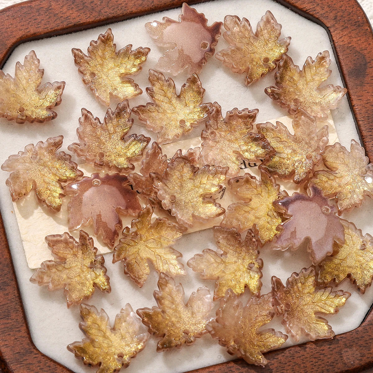 A collection of translucent amber and gold maple leaf glass beads arranged in a wooden tray.